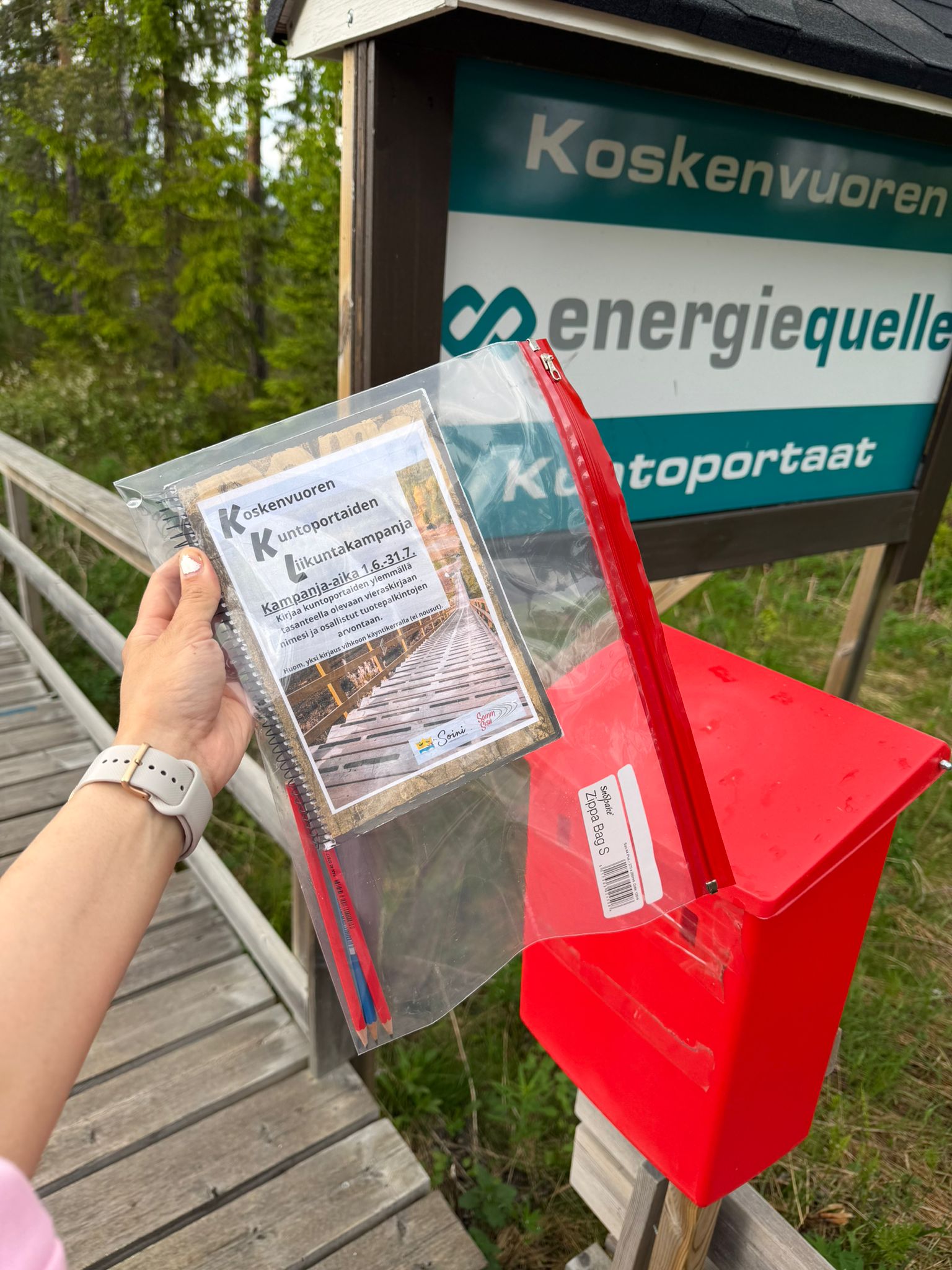Kuvassa on vihko muovitaskun sisällä ja taustalla punainen postilaatikko.